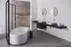 Badezimmeransicht mit schmaler Waschbeckenanordnung, zwei ovalen Waschbecken und runder Spiegelbeleuchtung. Freistehende weiße Badewanne und Dusche mit Glasschirm von Ideal Standard.